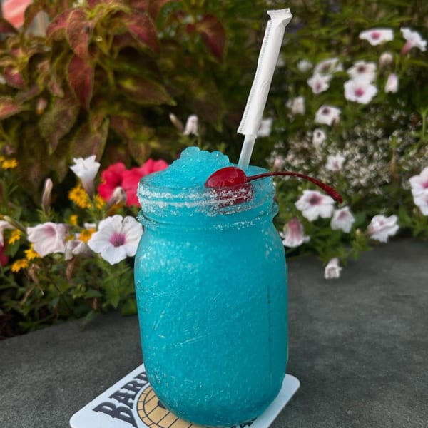 A vibrant blue frozen drink in a mason jar with a cherry and straw, set against a floral background.