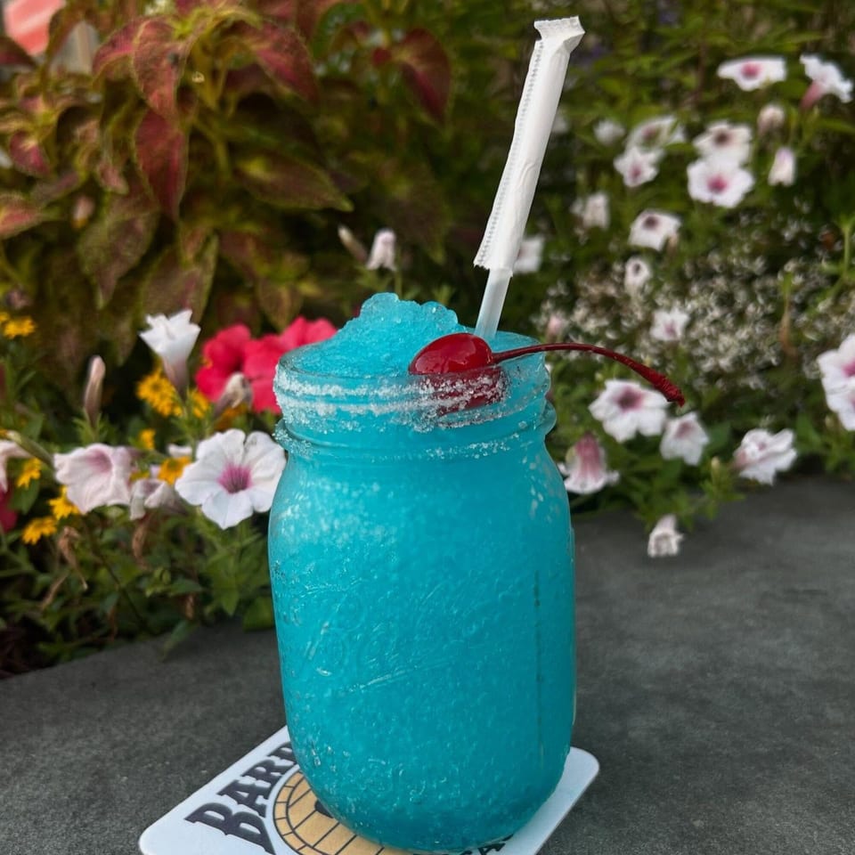 A vibrant blue frozen drink in a mason jar with a cherry and straw, set against a floral background.
