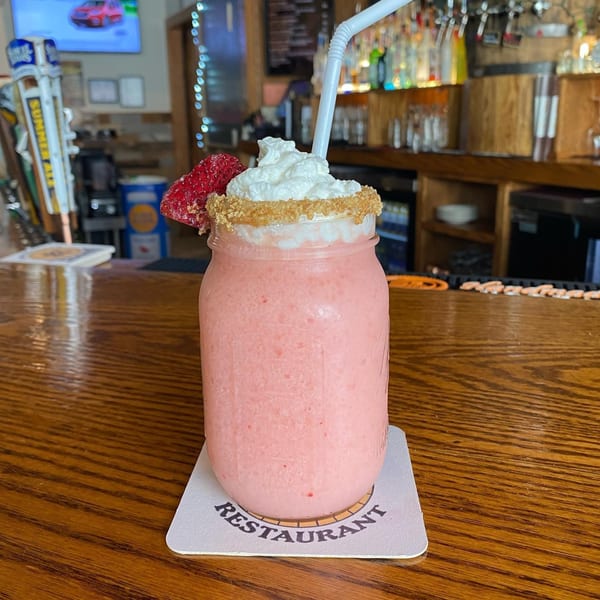 Pink strawberry milkshake in a mason jar with whipped cream and a strawberry on a bar top.
