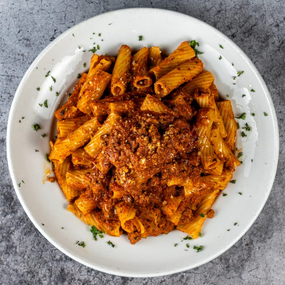 Rigatoni pasta with a rich red sauce, sprinkled with cheese and parsley, in a white bowl.