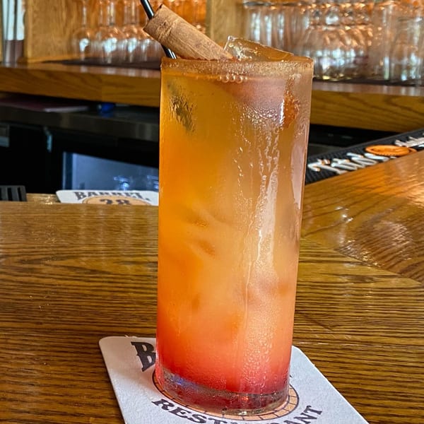 A tall, colorful cocktail with a cinnamon stick and sugared rim sits on a Barrel 28 coaster.