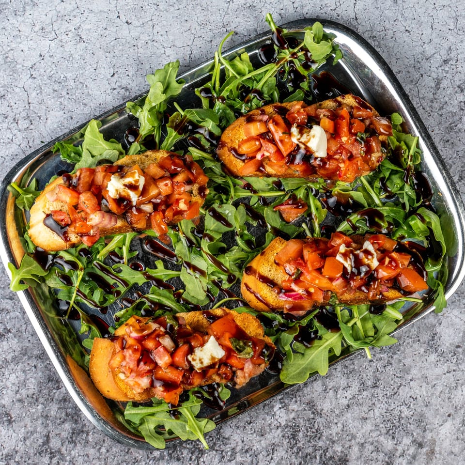 Four bruschetta pieces with diced tomatoes, cheese, and balsamic glaze on a bed of arugula.